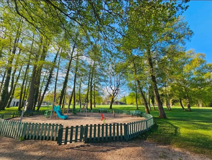 Aire de jeux clôturée sur sol sablé sous de grands arbres : toboggan turquoise, jeu à ressort et bancs.